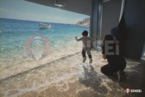 Un bambino salta tra le onde di una spiaggia proiettata sul pavimento mentre un'operatrice indica la barca sulla parete
