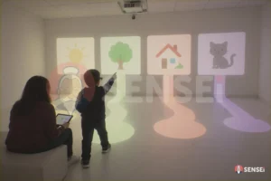 Un bambino con occhiali per il tracciamento dello sguardo sceglie un'immagine proiettata a parete, accanto a un'operatrice con tablet, alla Fondazione Angelo Custode