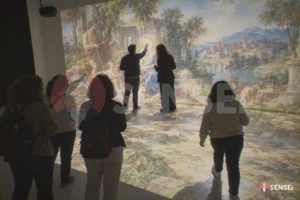 Un giovane mediatore museale tocca un dettaglio di un dipinto proiettato su parete e pavimento davanti a un gruppo di visitatori