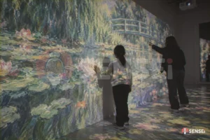 Una visitatrice tocca un paesaggio impressionista proiettato su parete e pavimento e fa sbocciare pennellate colorate, con una mediatrice museale
